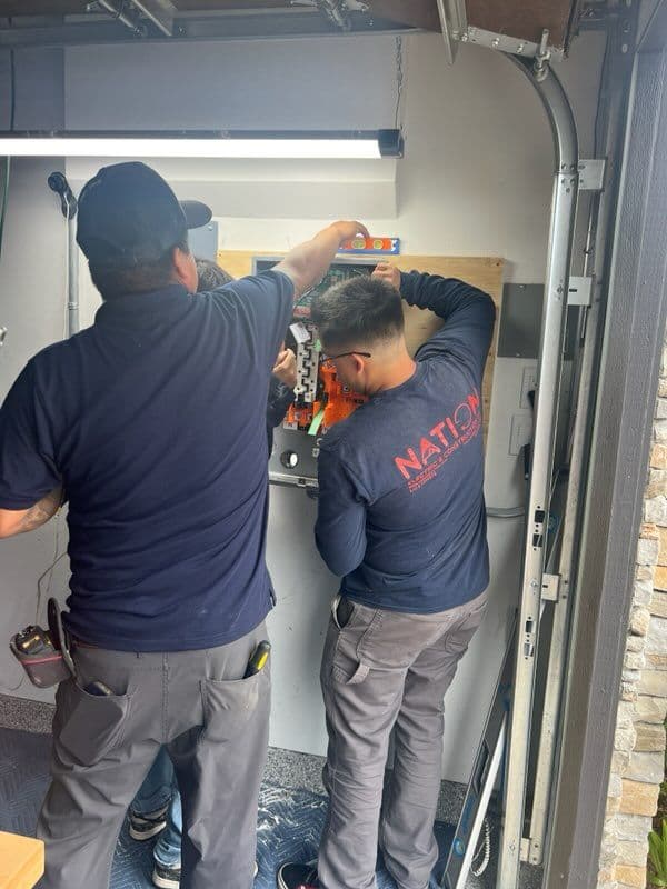 Two technicians work on an electrical panel in a well-lit garage environment.