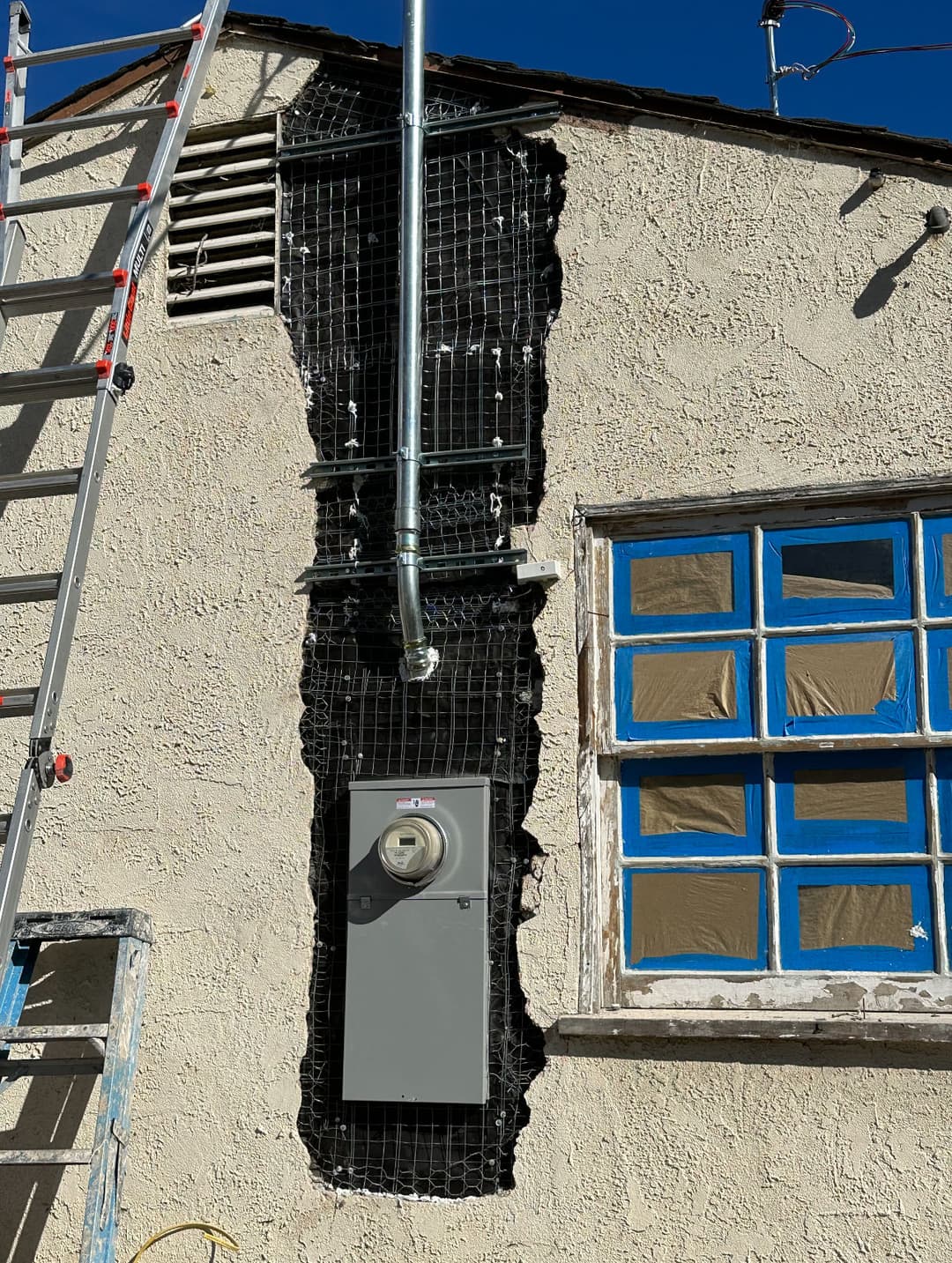 Ladder beside a wall with electrical meter and black netting for installation work.