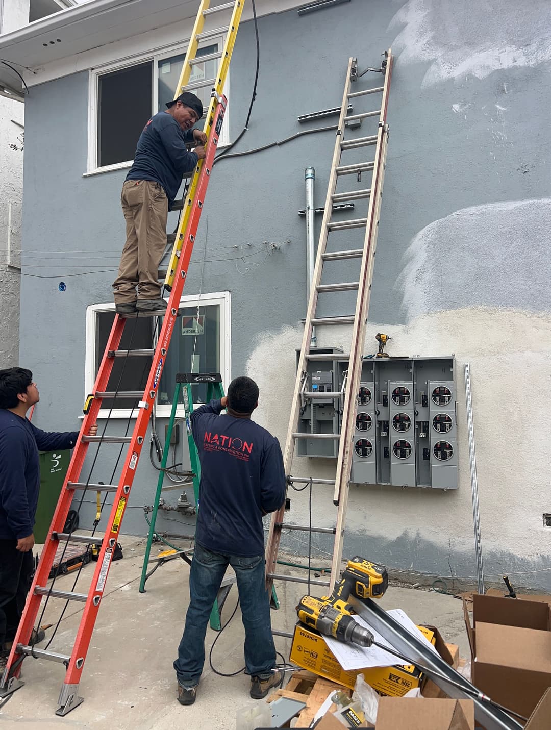 Workers installing electrical equipment on a building using ladders and tools.